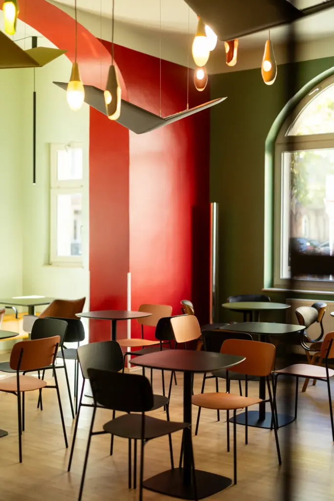 Innenraum des charc mit mehreren schwarzen Tischen, Stühlen in Schwarz, Terracotta und Braun, roter Wandfläche, olivgrünen Wänden und dekorativen Hängeleuchten