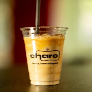 Eiskaffee in einem durchsichtigen To-go-Becher mit charc-Logo und dem Schriftzug „boards, brews & beyond", im Hintergrund unscharfer roter und grüner Farbverlauf